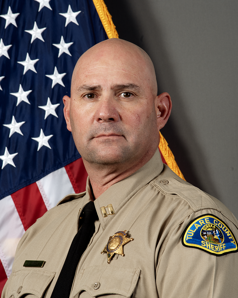A head and shoulders portrait of Captain Steve Sanchez III in uniform in front of an American Flag.