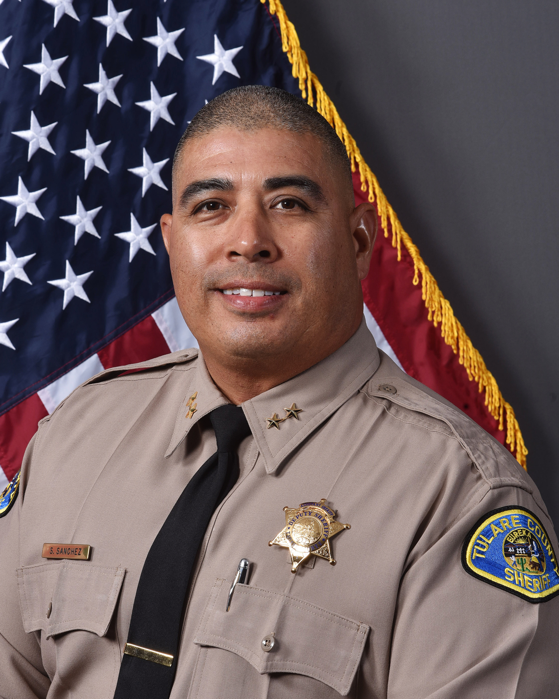 A head and shoulders portrait of Assistant Sheriff Steve Sanchez III in uniform in front of the American Flag.