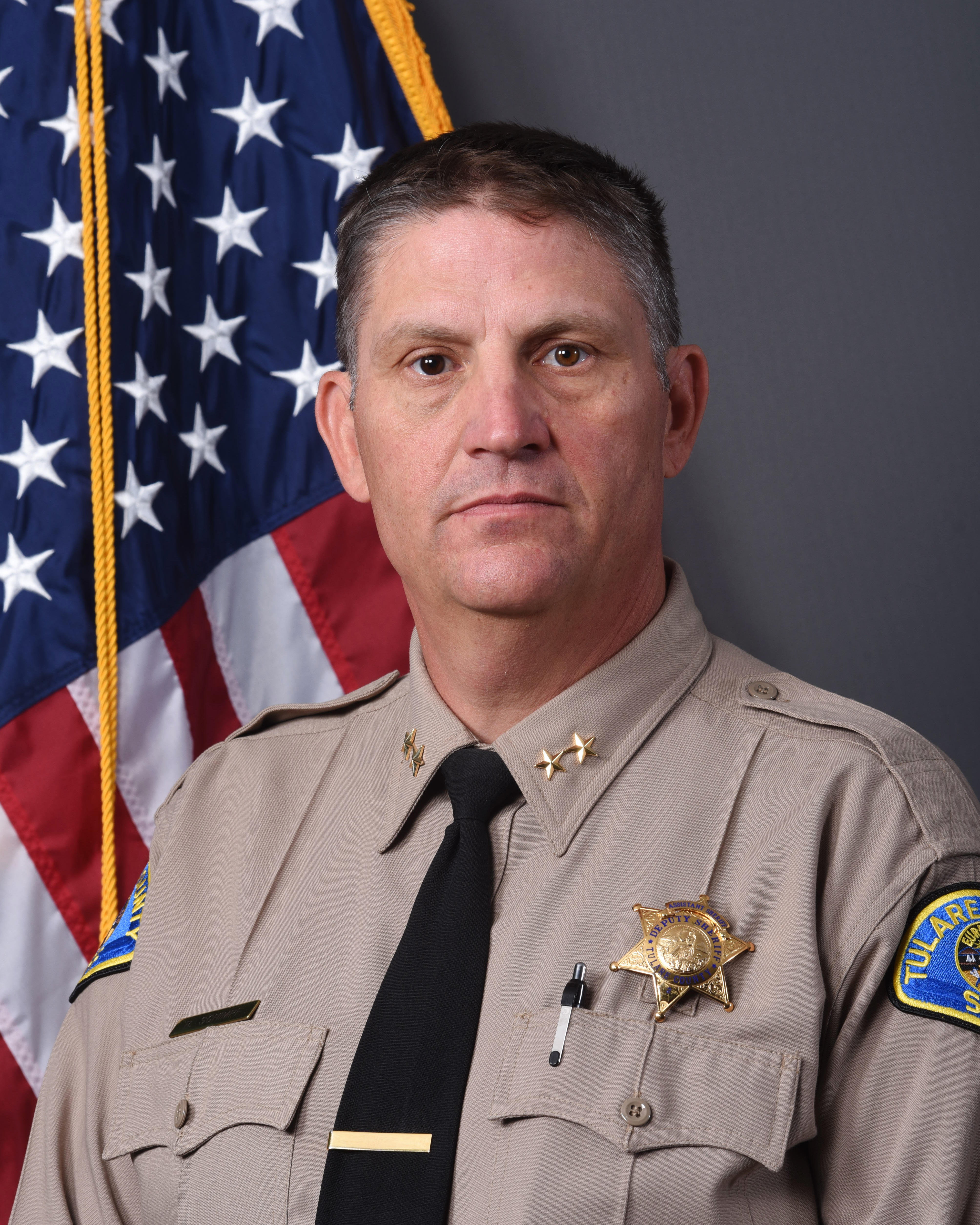 A head and shoulders portrait of Assistant Sheriff Robert Schimpf in uniform in front of the American Flag.