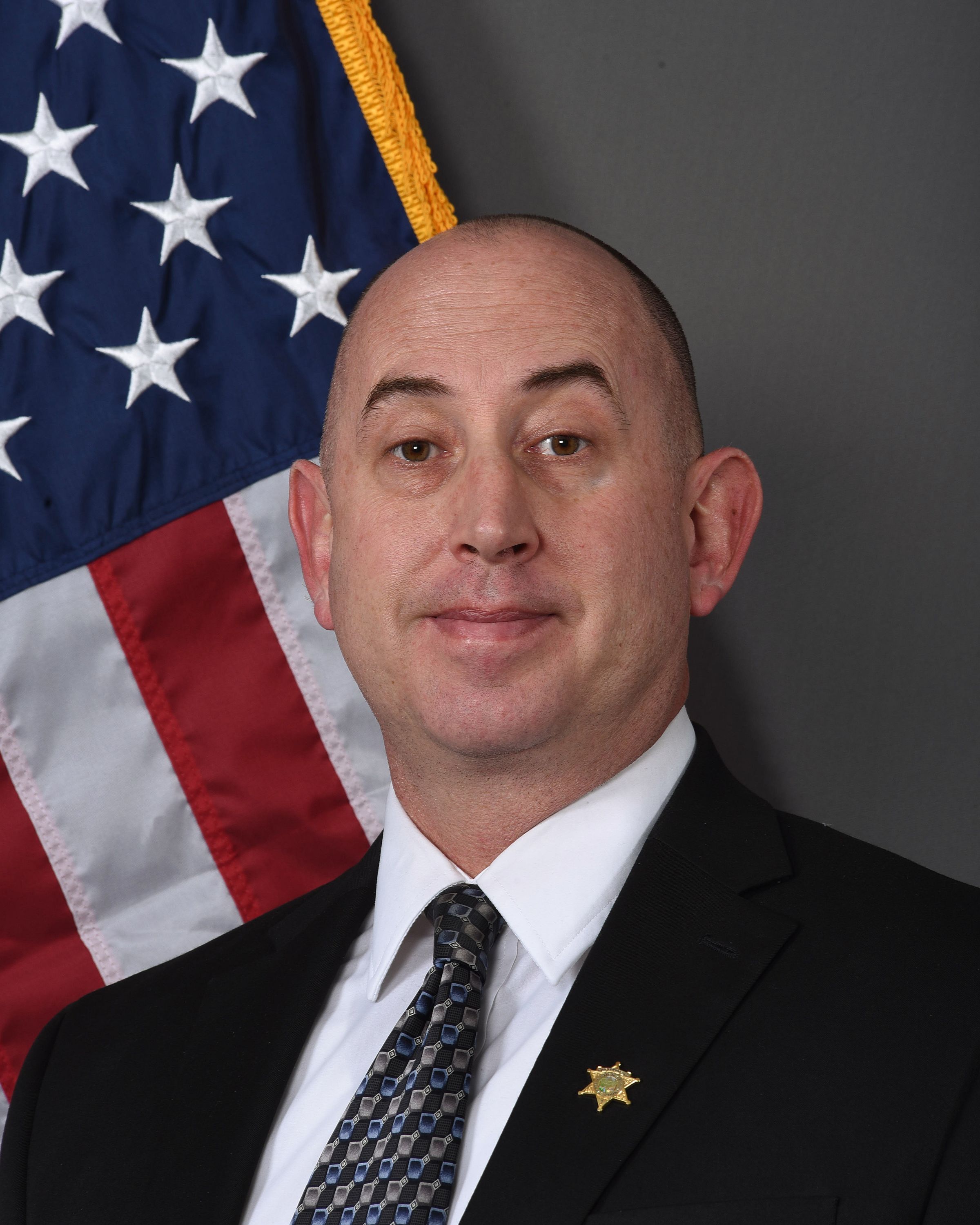 A head and shoulders portrait of Forrest McLean, Support Services Manager,  in a suit with the American Flag behind him. 