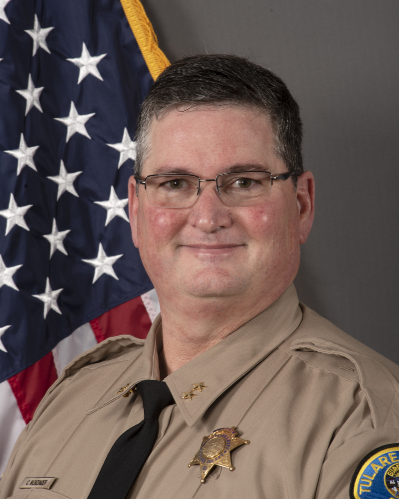 A head and shoulders portrait of Assistant Sheriff Chris Wenzinger in uniform in front of the American Flag.