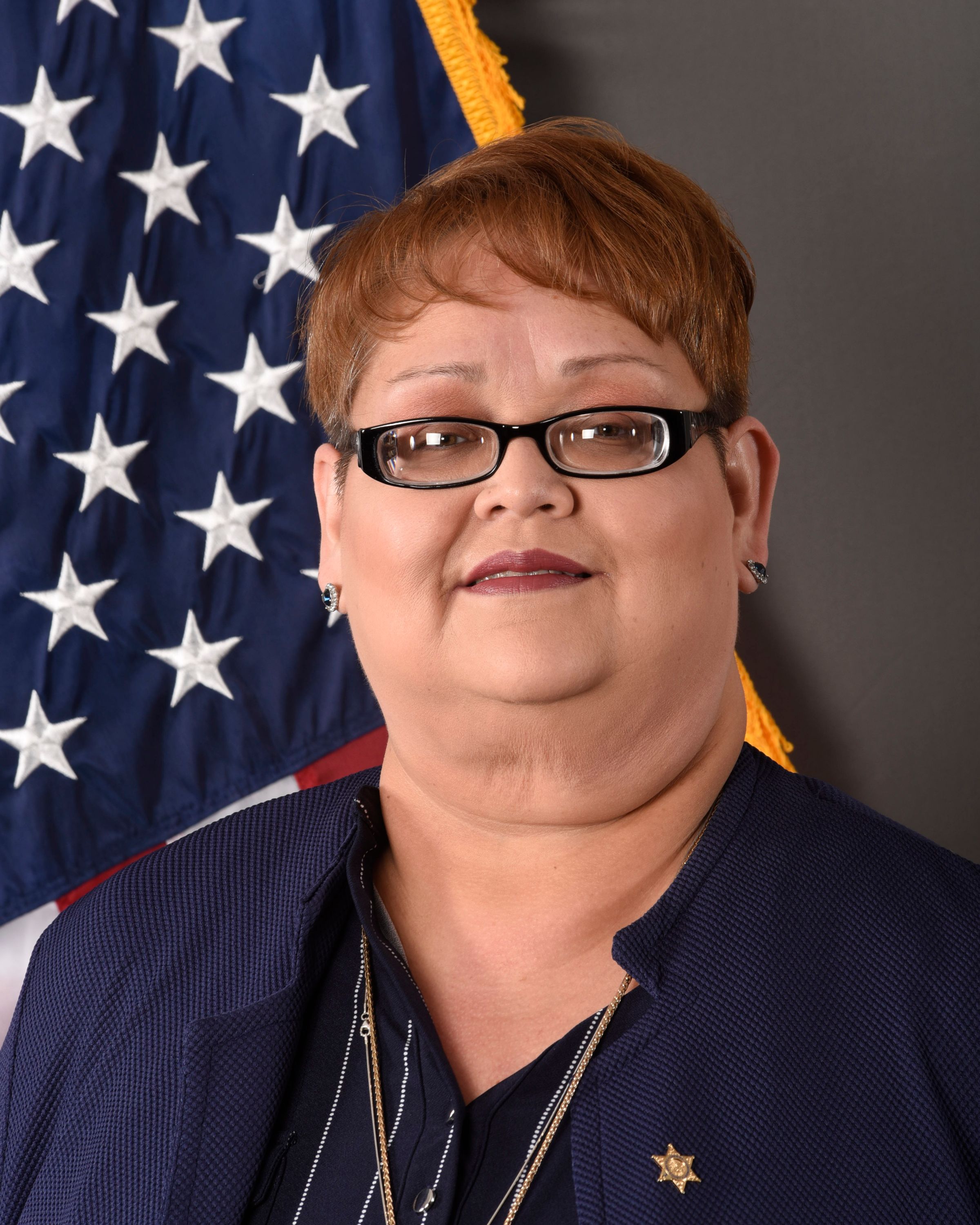 A head and shoulders portrait of Susanna Reyes, Inmate Programs Manager, in a navy blue collared blouse, standing in front of the American Flag.