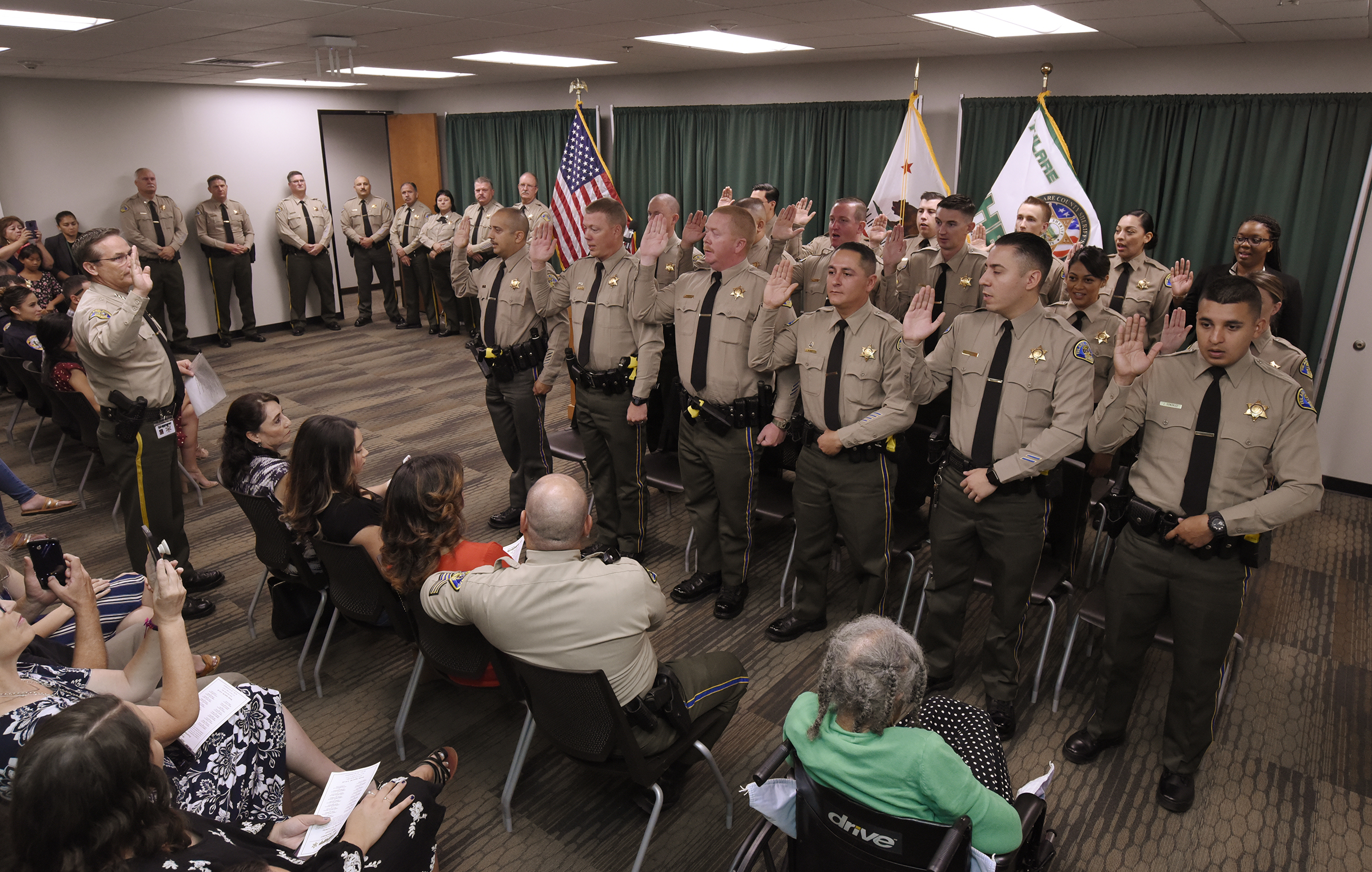 Sheriff Mike Boudreaux, pictured at left, with his right hand raised, swears in a large group of new deputies (three rows of uniformed men and women with their right hands raised) as a packed room of family, friends and colleagues look on.