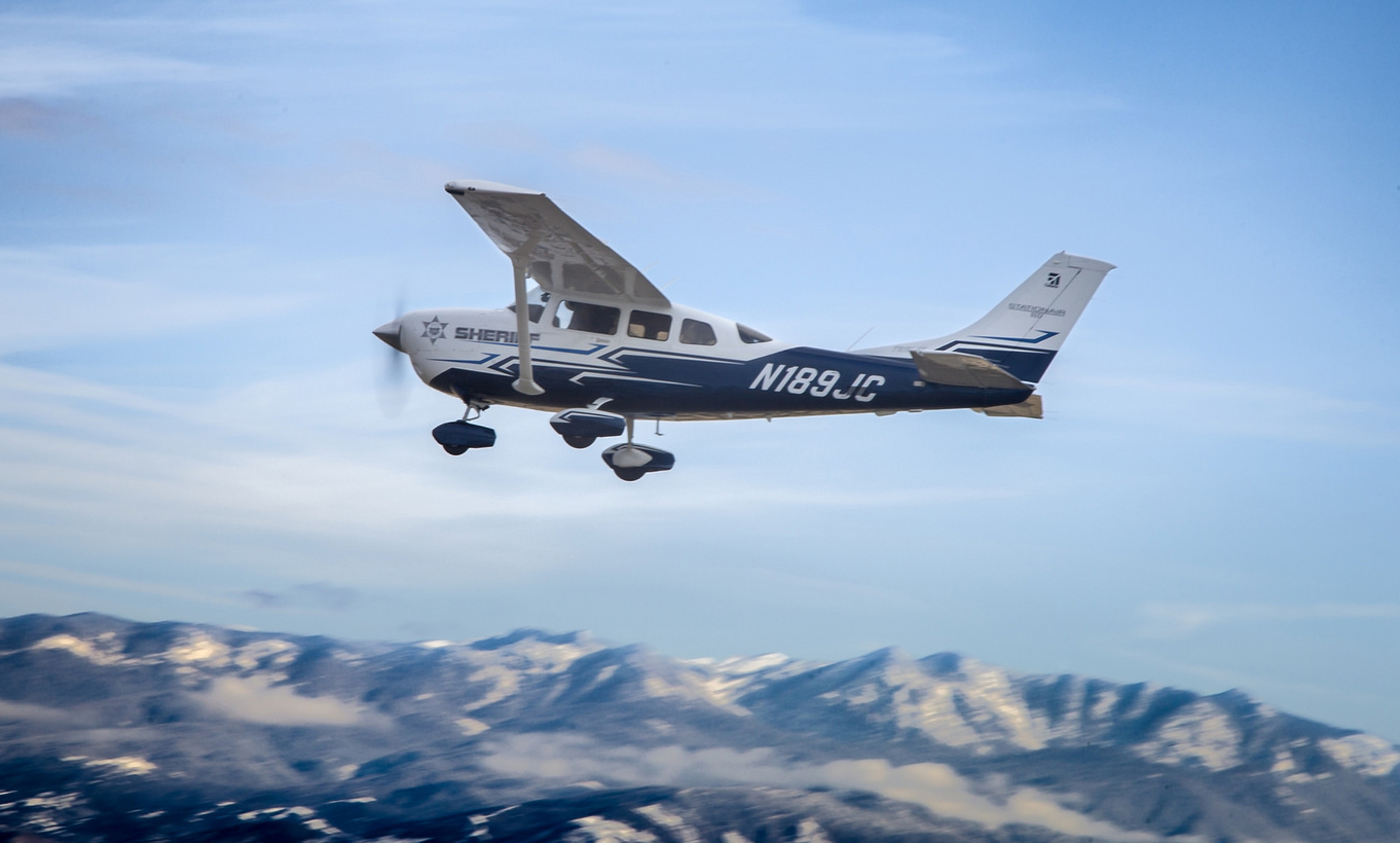 Tribute, the Sheriff's Cessna 206, can be see from another airplane flying a little below Tribute. Behind Tribue is the blue sky with some white clouds visible and below the plane is the Sierra Nevada mountains. The call sign N189JC is visible in white lettering on blue toward the tail.