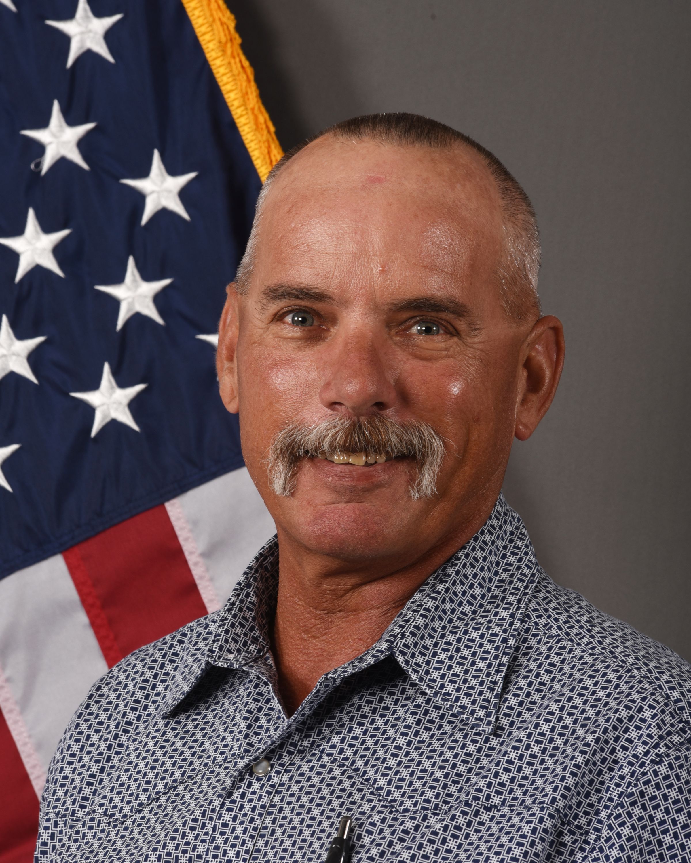 A head and shoulders portrait of Gary Bird, Farm Manager, in a western style shirt in front of the American Flag.