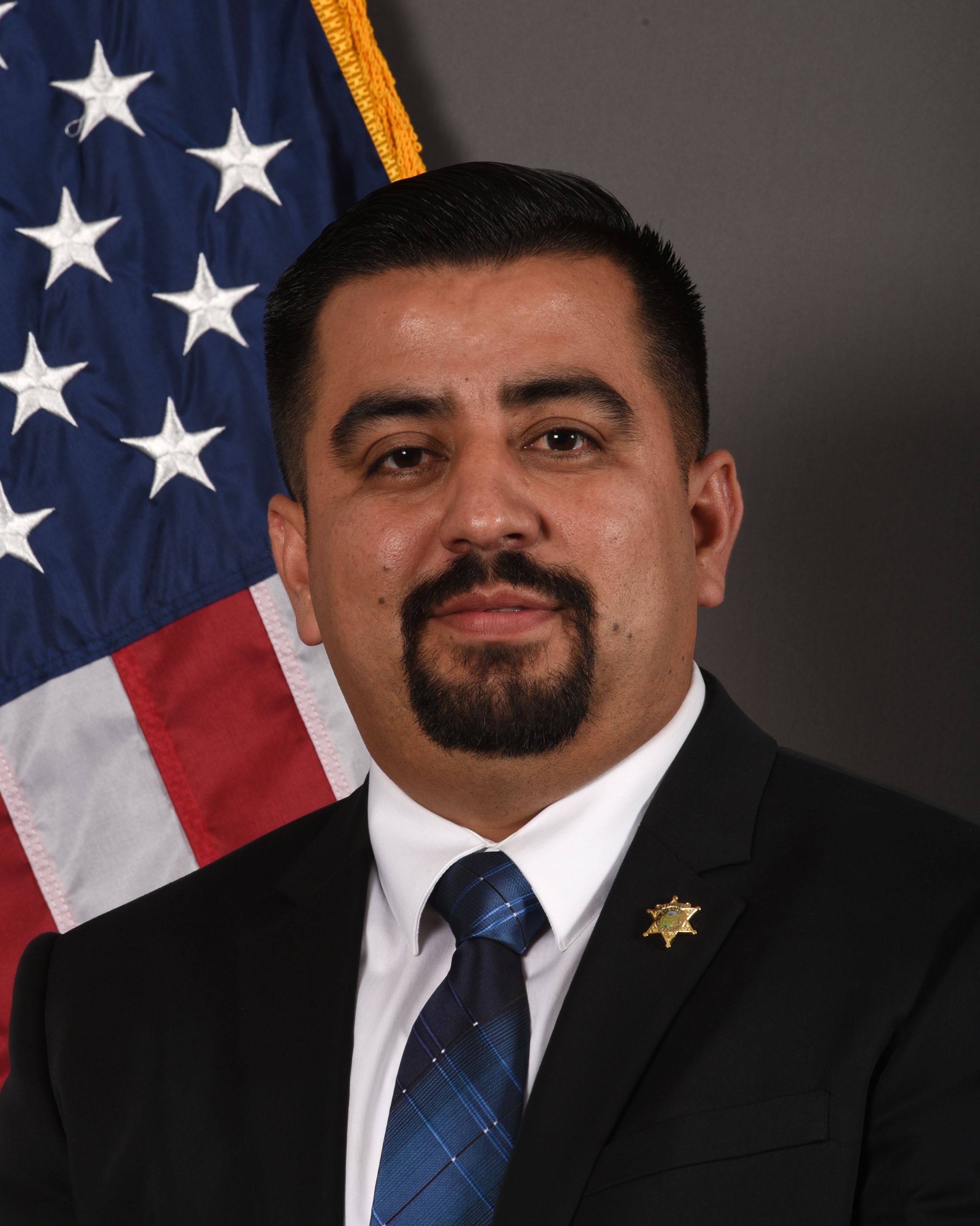 A head and shoulders portrait of Alfredo Parra, Fiscal Manager, in a suit in front of the American Flag. 
